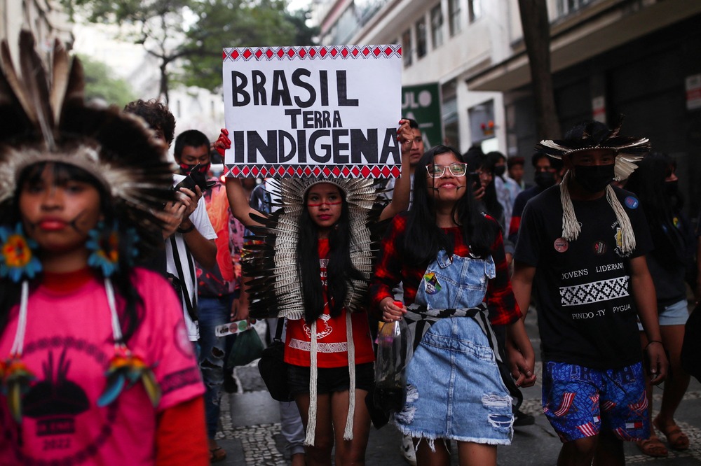 People wearing indigenous garb and ornamentation march in street; one person holds sign reading 'Brazil is Indigenous Land'.