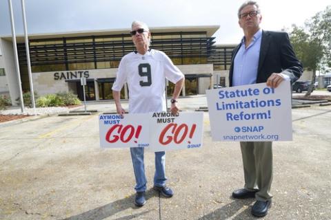 Two men stand outside of New Orleans Saints building, holding signs calling for Archbishop Gregory Aymond's dismissal,  and statute of limitations reform. 
