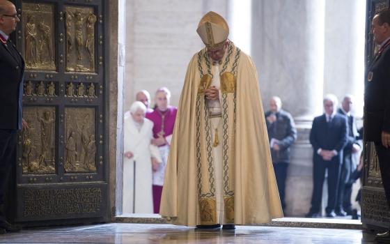 Pope Francis, vested, walks with head bowed through Holy Doors of St. Peters; in background is Pope Emeritus Benedict XVI, wearing plain white coat, supported by cane and bishop. 