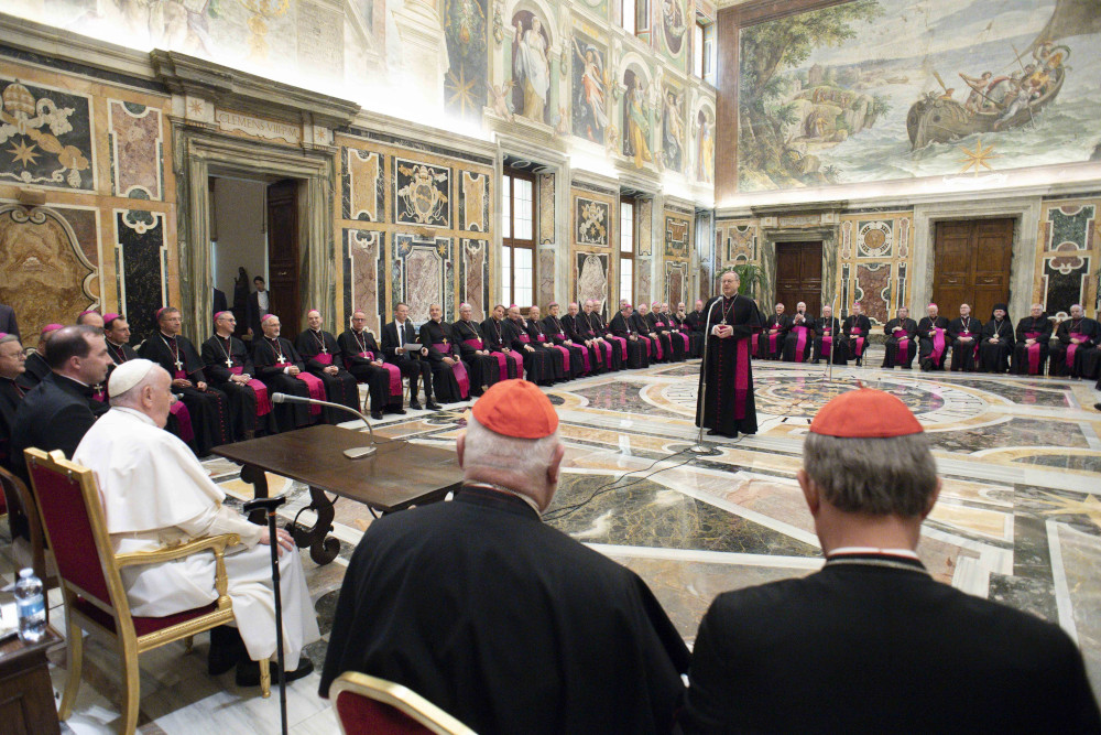A bishop stands in the middle of an ornate room and speaks to Pope Francis and some cardinals