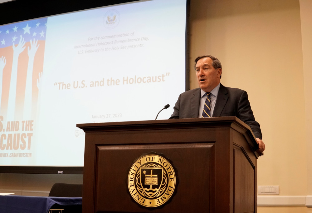 A white man in a suit speaks from behind a podium with a projector and the words "The U.S. and the Holocaust" in the background