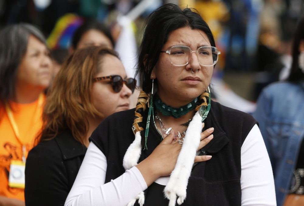 A woman with long beaded earrings and glasses holds a hand to her chest. Her braids are wrapped in leather