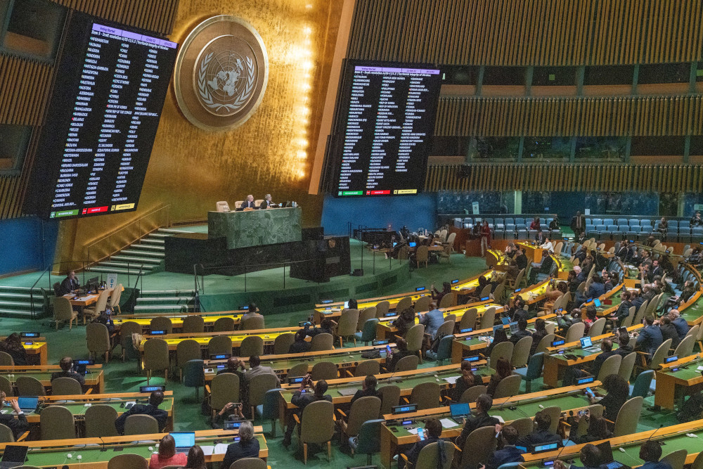 Semicircles of long desks with chairs and people with a raised dais with large screens and the UN symbol