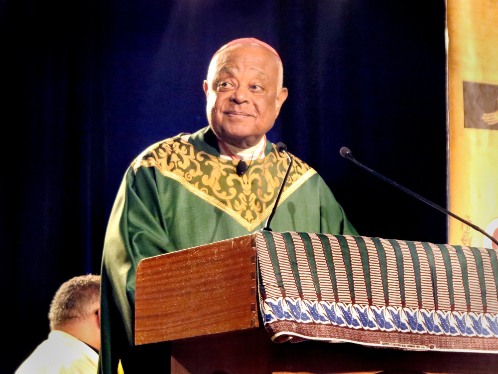 A Black bald man wears green and gold vestments and speaks from behind a podium