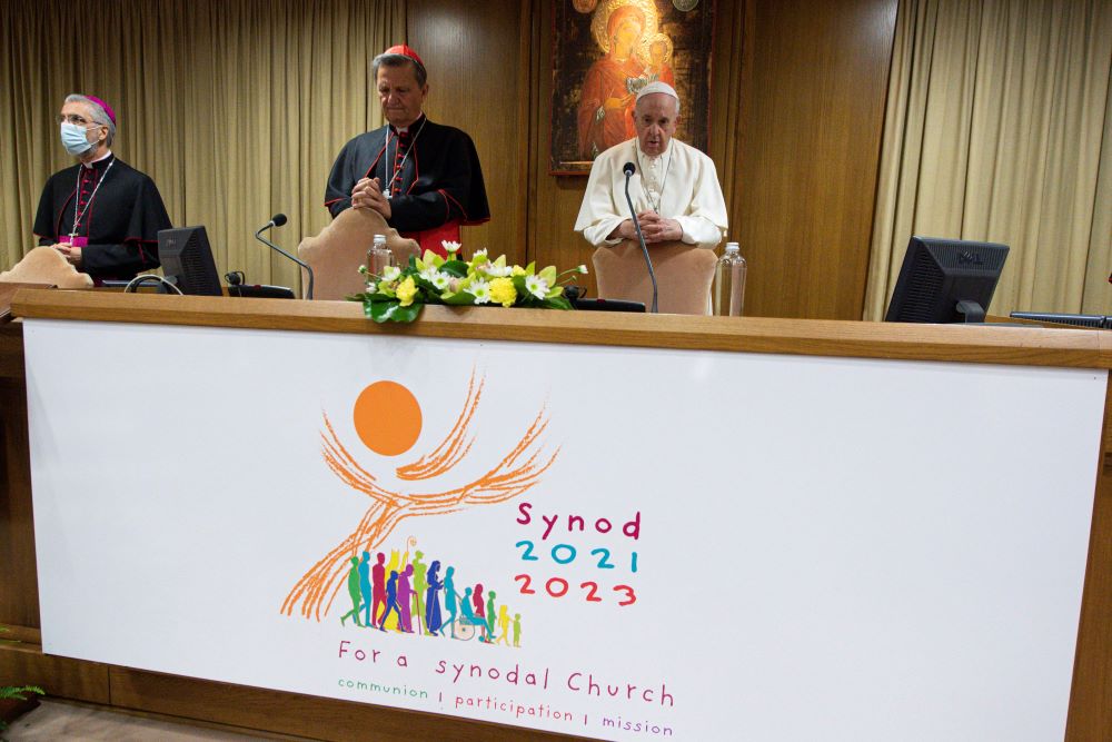 Vatican officials, including Cardinal Grech and Pope Francis stand at table with large banner with synod logo