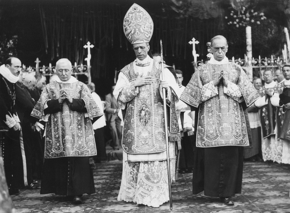 A black and white photo with a man in the middle wearing a mitre and carrying a crozier surrounded by other men in vestments