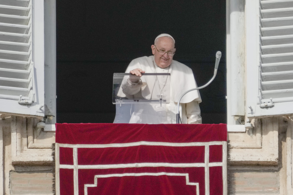 Pope Francis smiles and peers over the edge of his apartment window with one hand on his clear lectern 