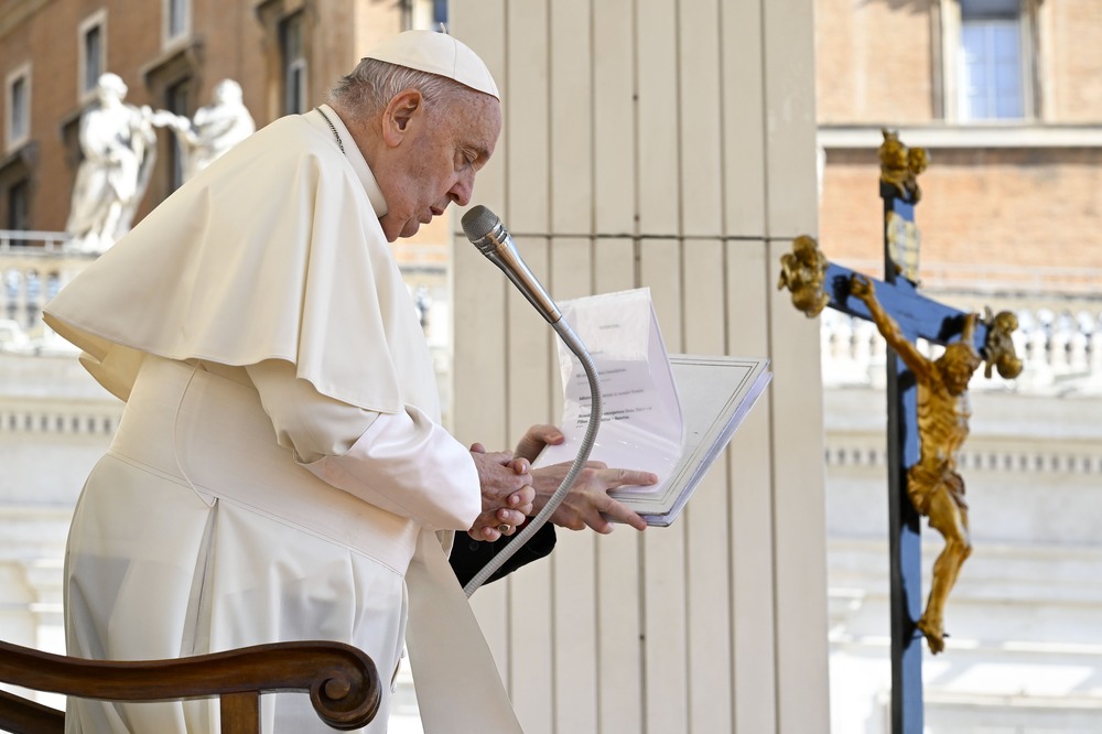 Pope Francis stands, reading from notes, large crucifix hangs in background 