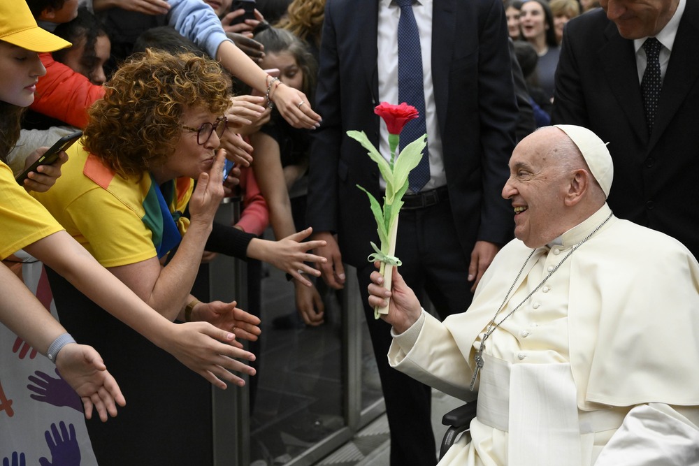 Pope Francis, in wheelchair, holds flower, and smiles at woman who blows kiss from behind barrier.