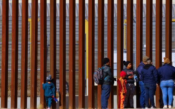 Adults and one small child line up along a towering fence of rust-colored metal posts