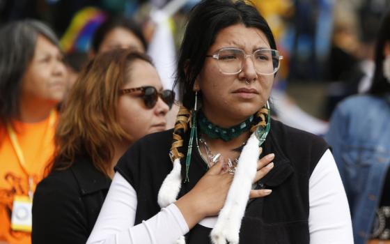 A woman with long beaded earrings and glasses holds a hand to her chest. Her braids are wrapped in leather