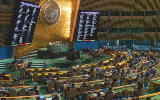 Semicircles of long desks with chairs and people with a raised dais with large screens and the UN symbol
