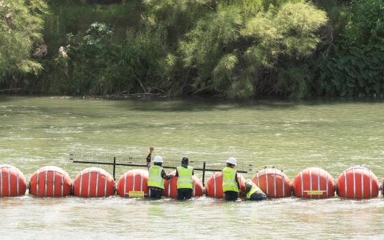 Three men in bright yellow vests address large orange buoys spaced closely in the river