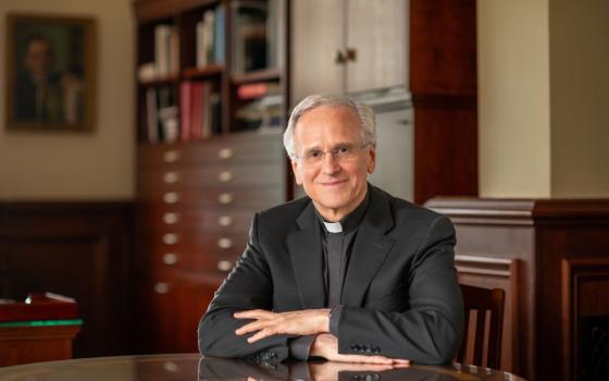 An older white man wears a clerical collar and black suit jacket in an office space