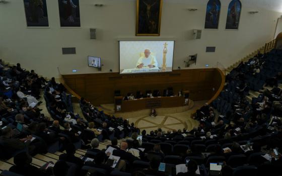 Pope Francis seen on screen in front of large seated assembly. 