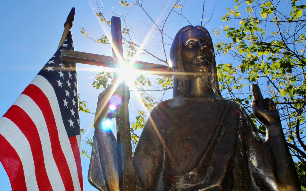 Sun shines through a statue of Christ on a grave marker alongside an American flag at St. Mary Catholic Cemetery in Appleton, Wisconsin. (CNS/Bradley Birkholz)