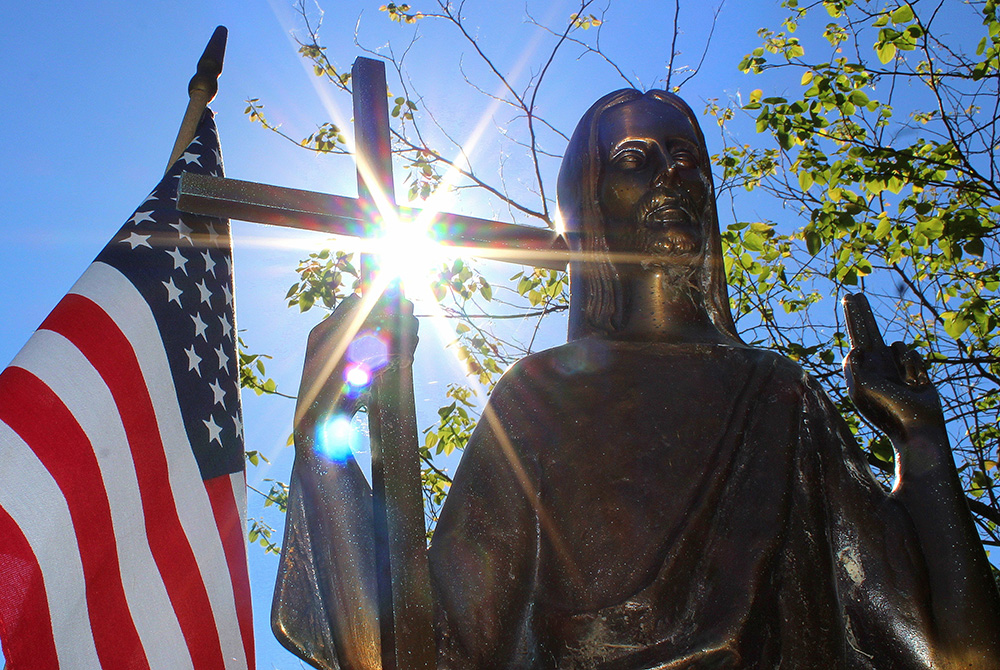 Sun shines through a statue of Christ on a grave marker alongside an American flag June 7, 2018, at St. Mary Catholic Cemetery in Appleton, Wisconsin. (CNS/Bradley Birkholz)