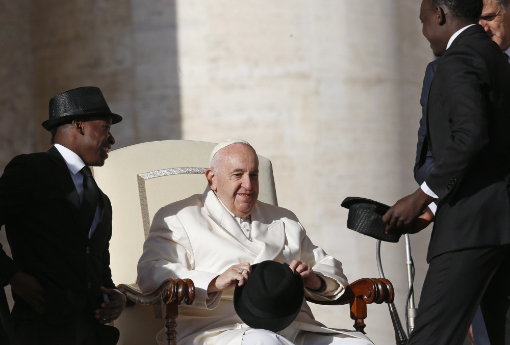 Pope Francis sits in a white chair and meets three Kenyan acrobats in top hats and black suits