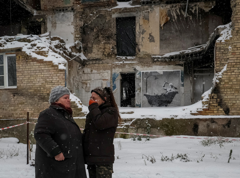 Two women stand outside a crumbling building in the snow. One woman cries.