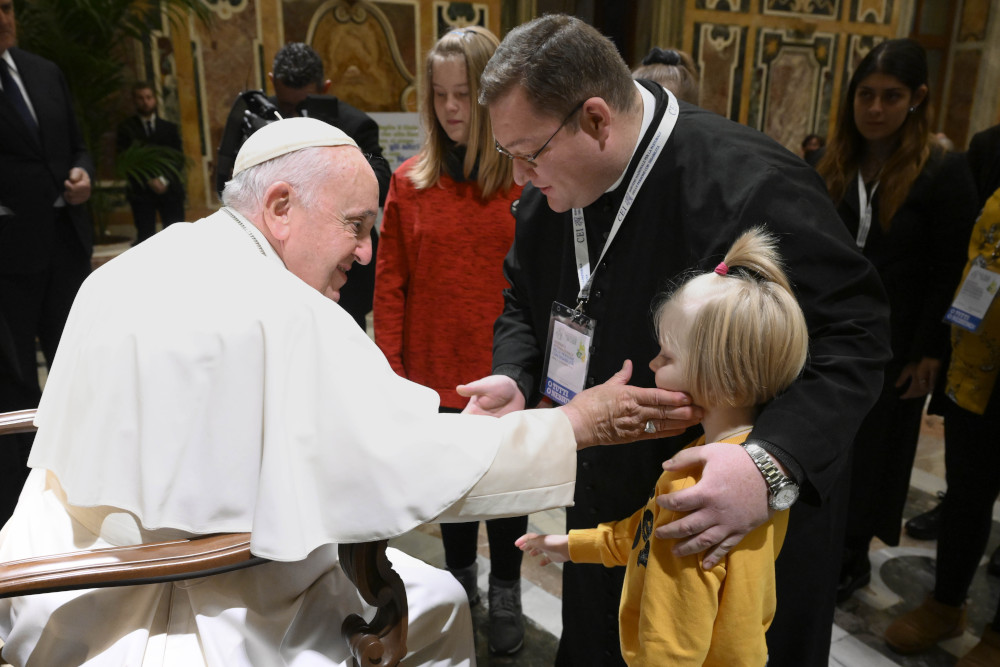 Pope Francis sits in a chair and touches a child's cheek as the child's father stands beside them