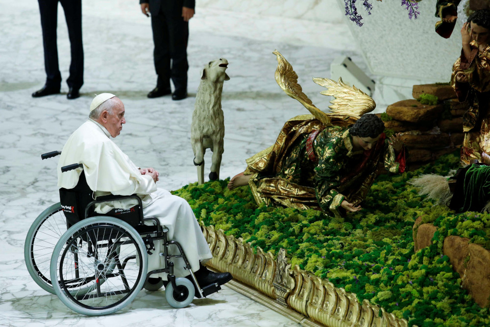 Pope Francis sits in a wheelchair in the snow facing a brightly painted creche