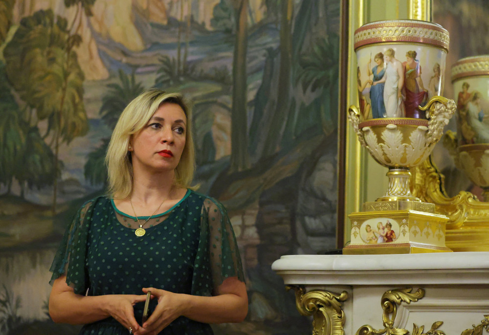 A blond woman with dark roots stands in front of a painting and ornate vase