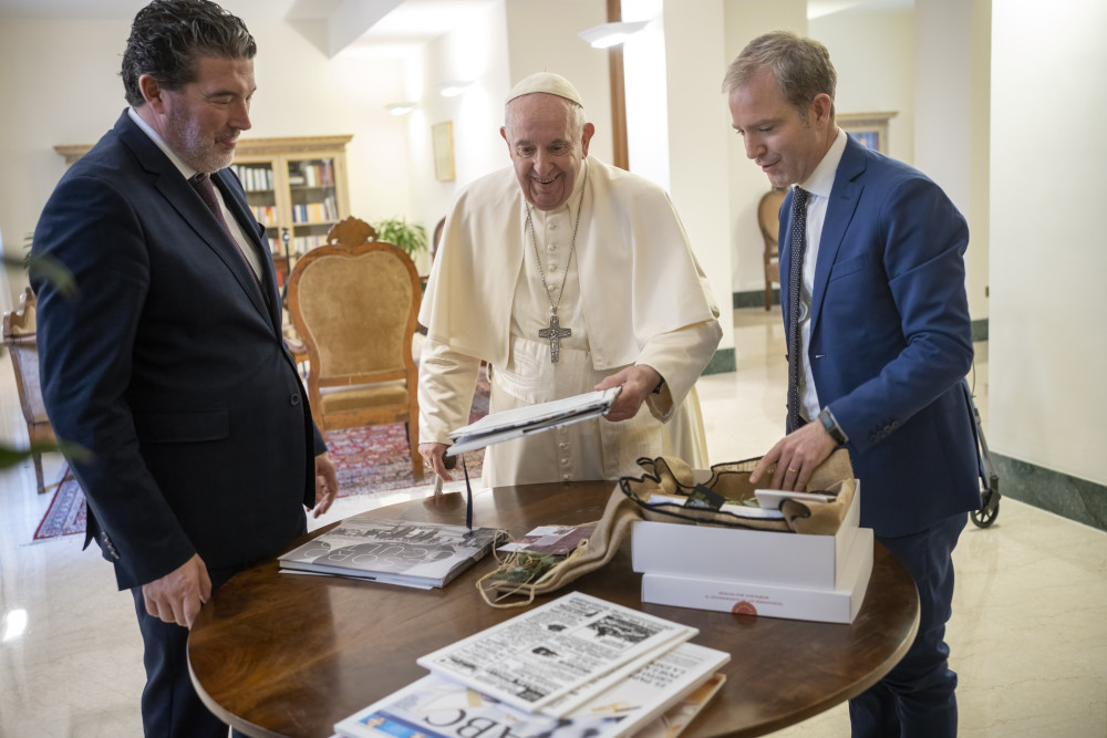Pope Francis and two Spanish men stand around a table with newspapers and books on it