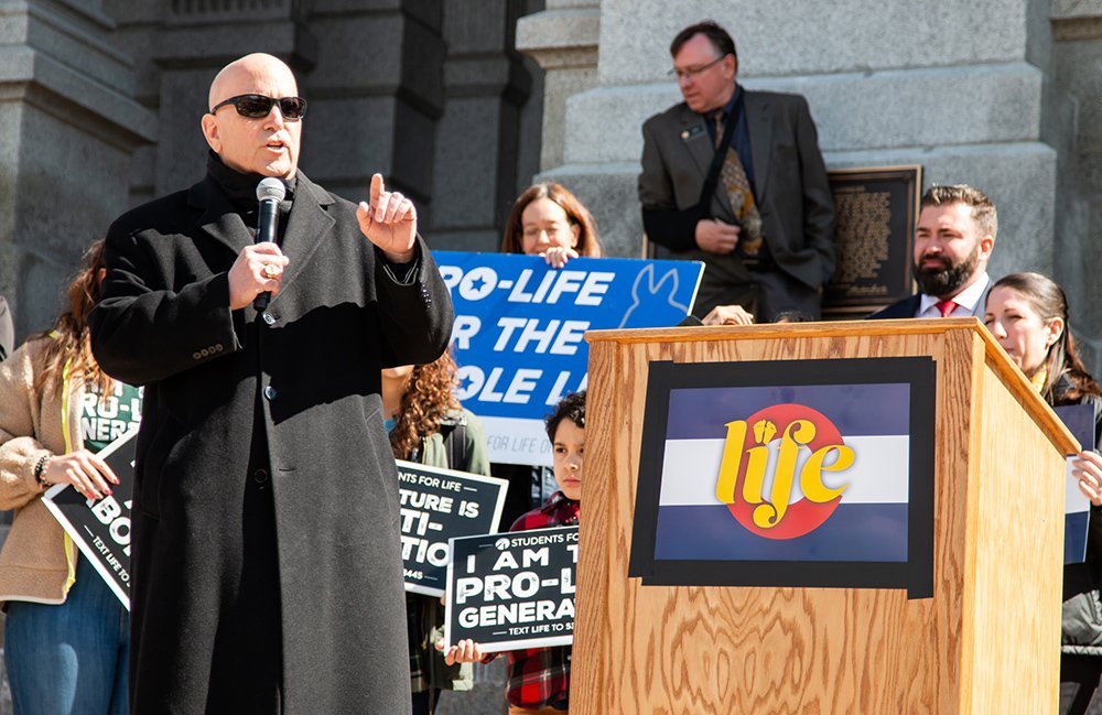 A bald man in black sunglasses and a long black coat holds a microphone and speaks in front of a crowd holding pro-life signs