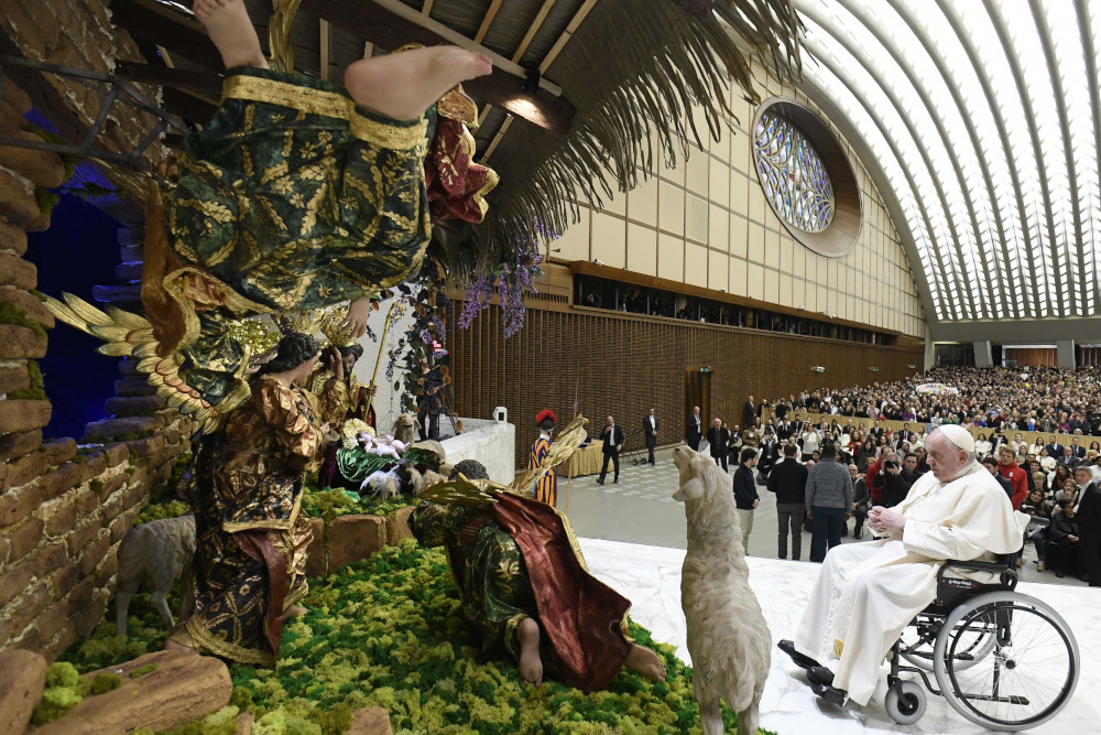 Pope Francis sits in a wheelchair in front of a large nativity scene indoors
