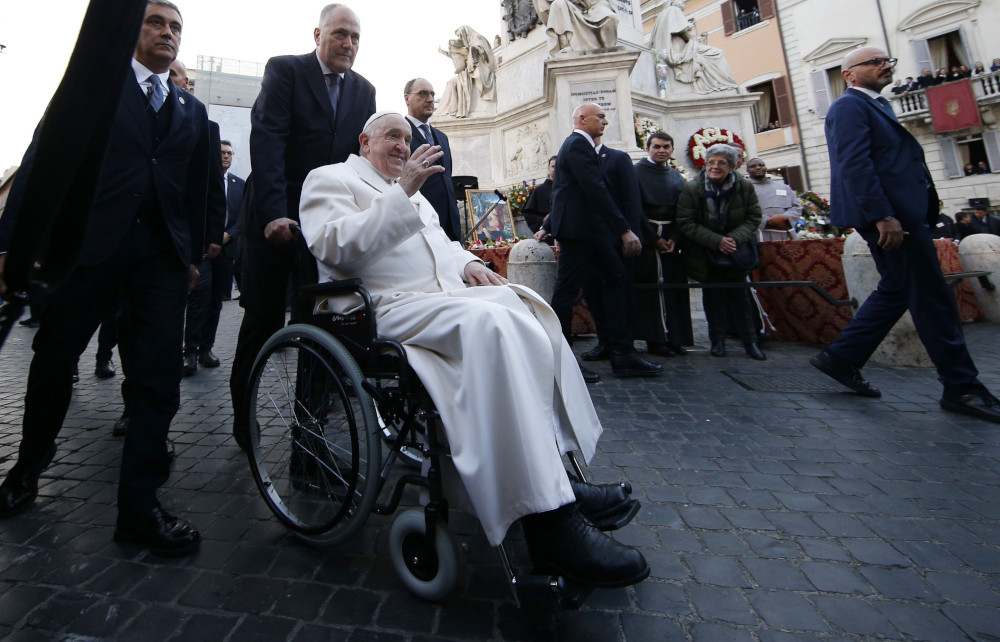 Pope Francis sits in a wheelchair pushed by a man in a suit and waves near a variety of statues