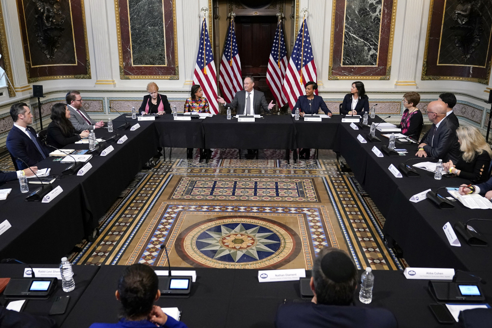 People sit at tables in a rectangle with Dough Emhoff at one end flanked by several American flags