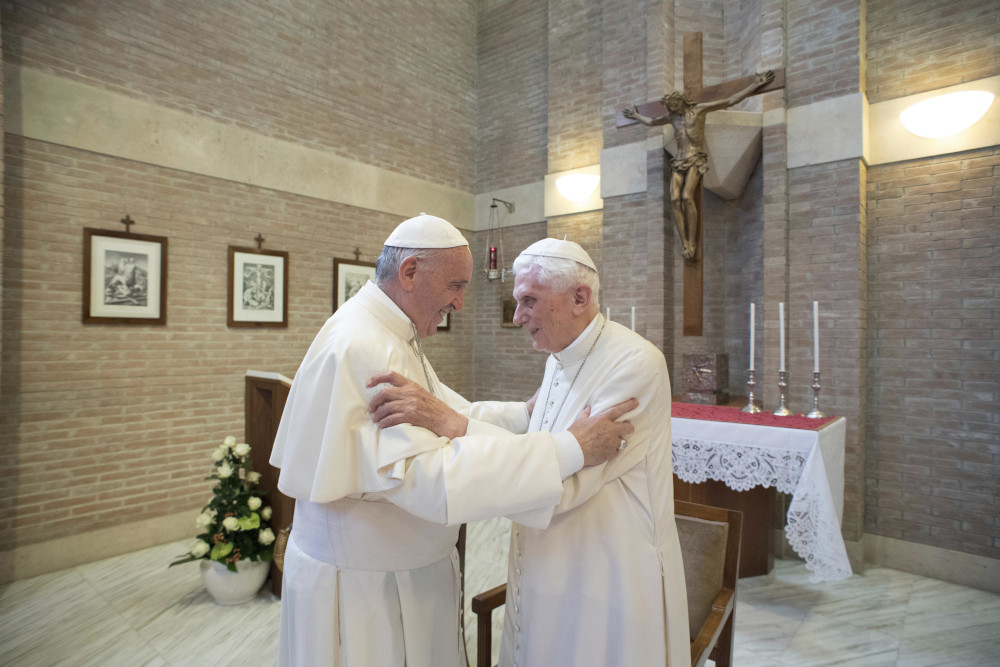 Pope Francis and Pope Benedict XVI, both in white cassocks, grip each others arms in a room with a crucifix and altar