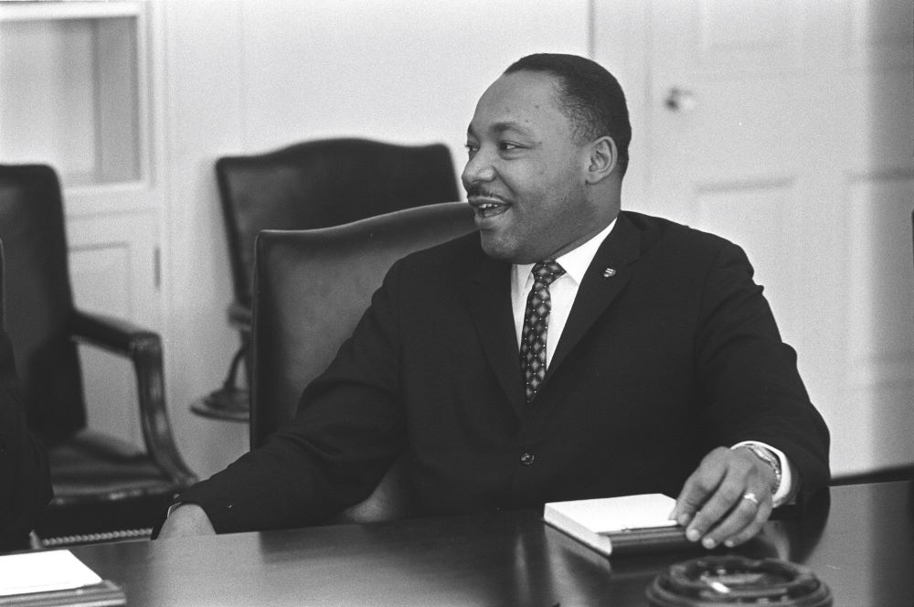 The Rev. Martin Luther King Jr. smiles while sitting at a table and talking.