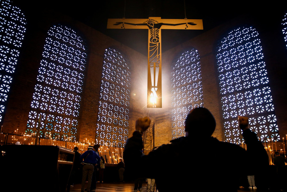 A person in shadow raises both fists below an illuminated crucifix