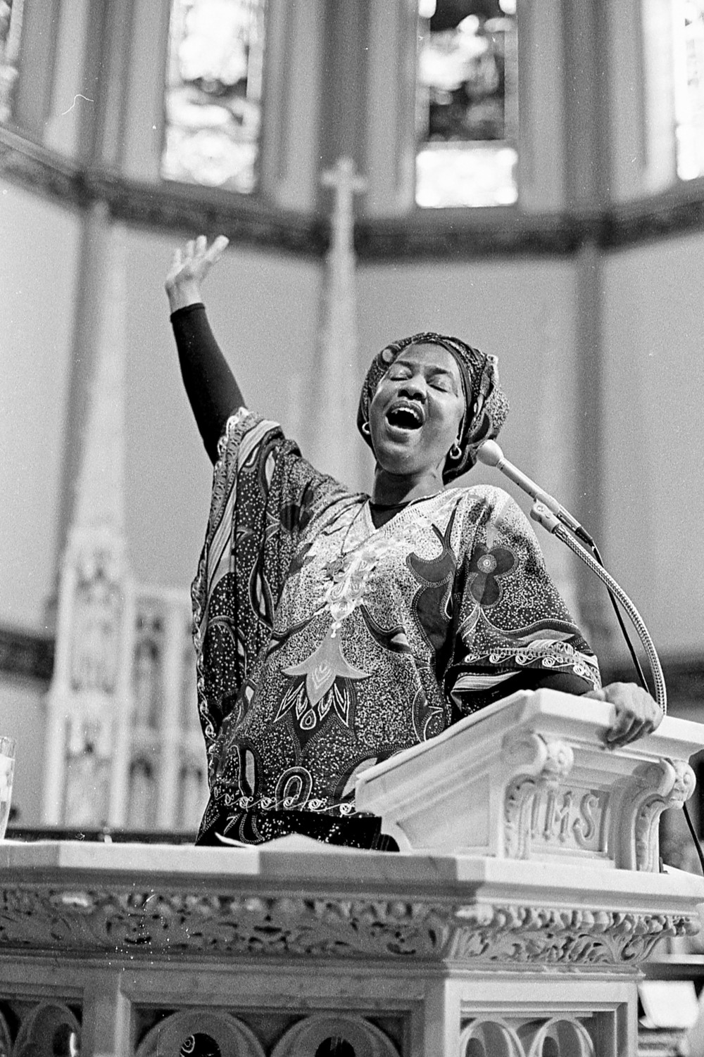 A black and white photo of a Black woman at a lecturn with one arm raised in the air. She wears patterned fabric around her head and torso.