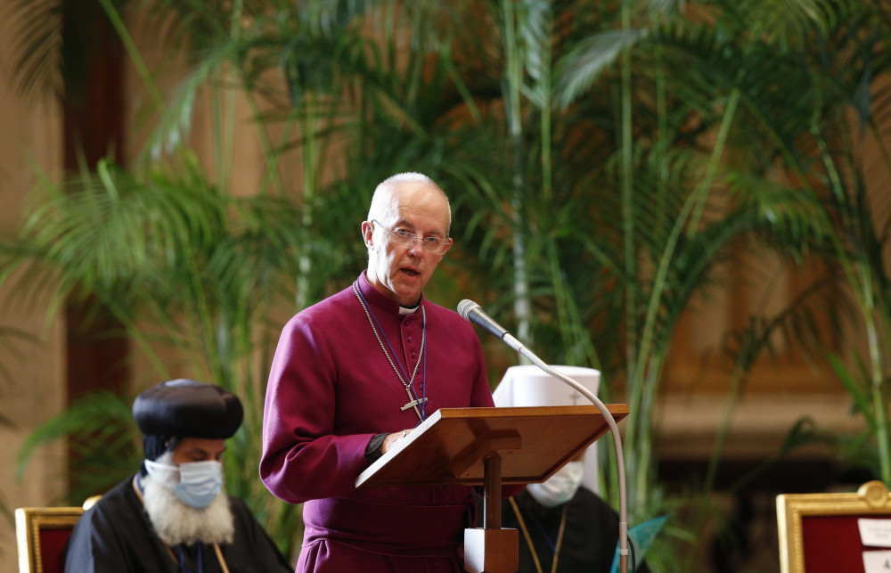 Justin Welby, an older white man wearing glasses, a cross, and a magenta clergy shirt, speaks into a microphone with large plants behind him