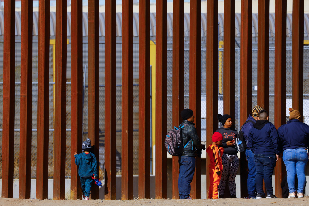 Adults and one small child line up along a towering fence of rust-colored metal posts