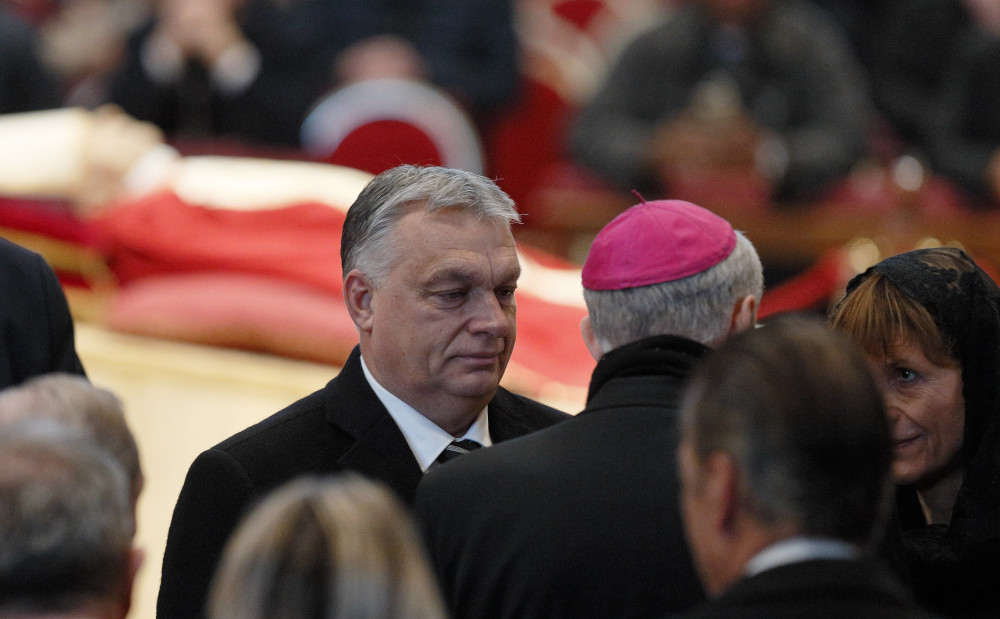 A gray-haired man in a suit talks to a bishop seen from the back with Pope Benedict XVI's body in the background