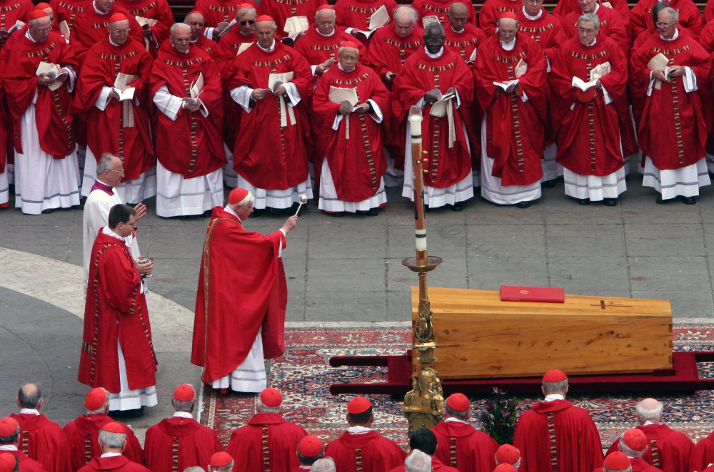 Many cardinals wearing red surround a wooden casket on a rug in St. Peter's Square