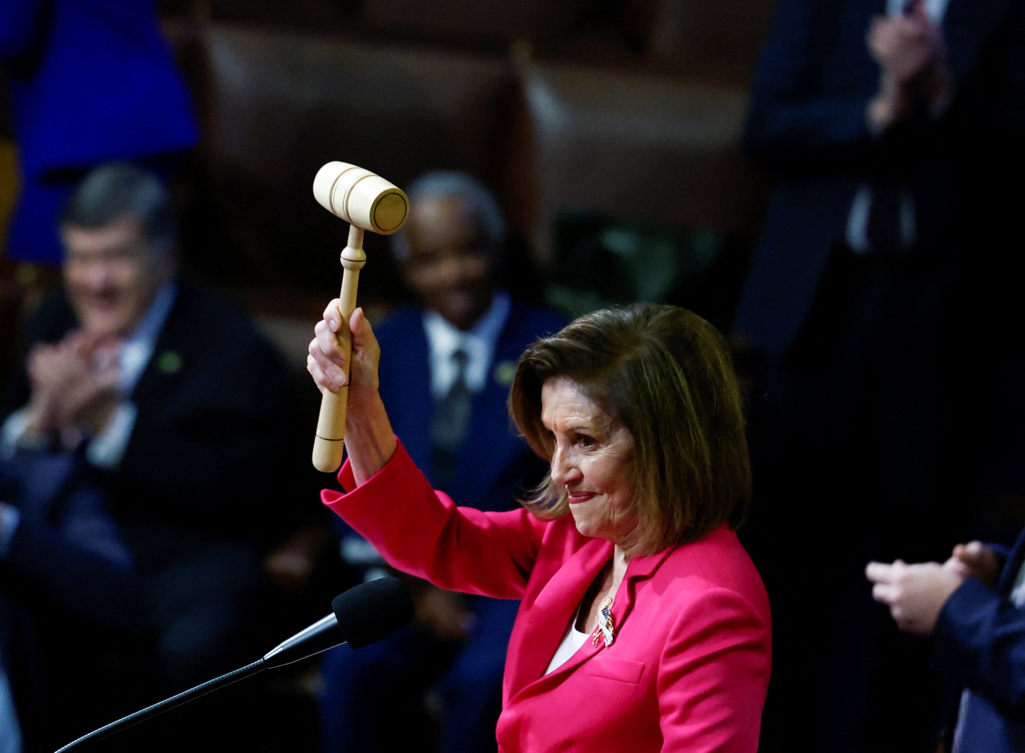 Nancy Pelosi, wearing a bright pink blazer, lifts a gavel high in the air