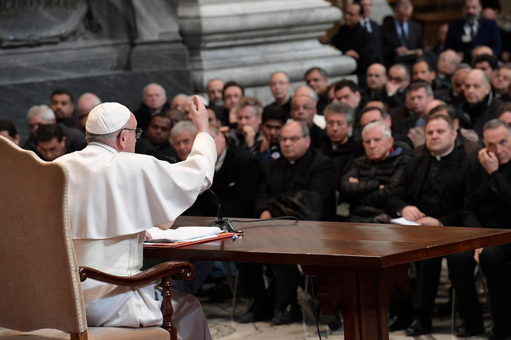 Pope Francis sits at a large table in front of a rows of many priests 