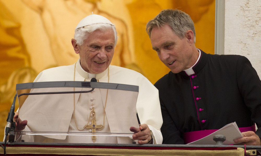 A white archbishop stands beside and leans toward Pope Benedict XVI