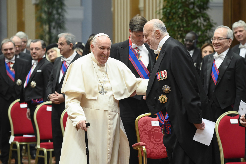 Pope Francis smiles broadly, holds a cane, and pats the arm of a white man wearing a tuxedo