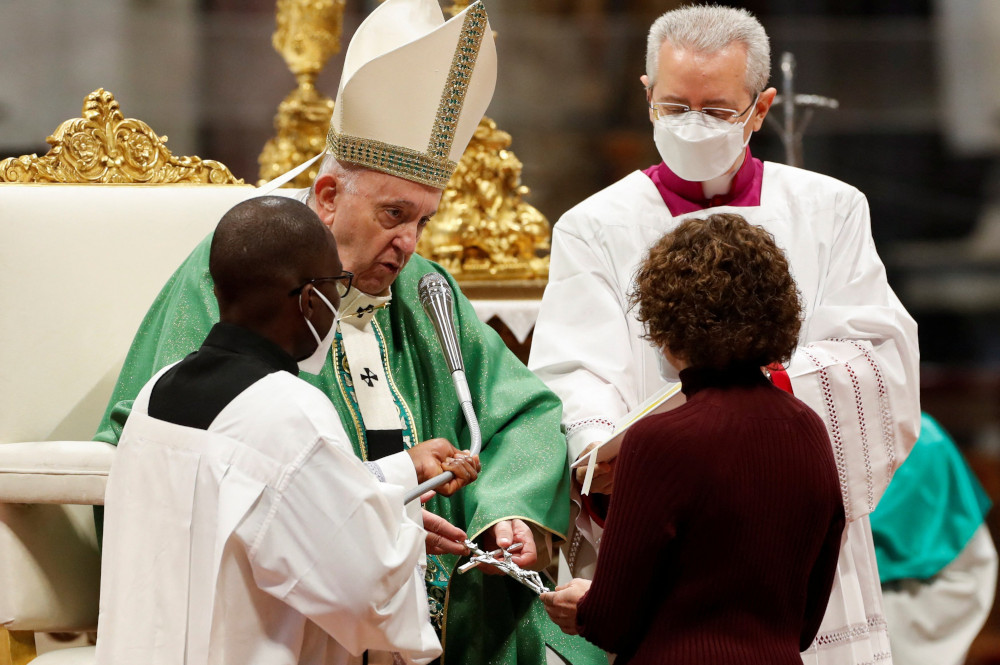 Pope Francis, wearing a mitre and no mask, is flanked by two masked priests and hands a crucifix to a masked woman