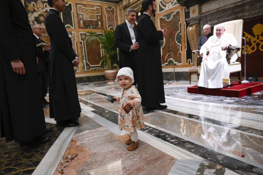 A toddler with a white hat stands in front of men in cassocks who wait to greet Pope Francis