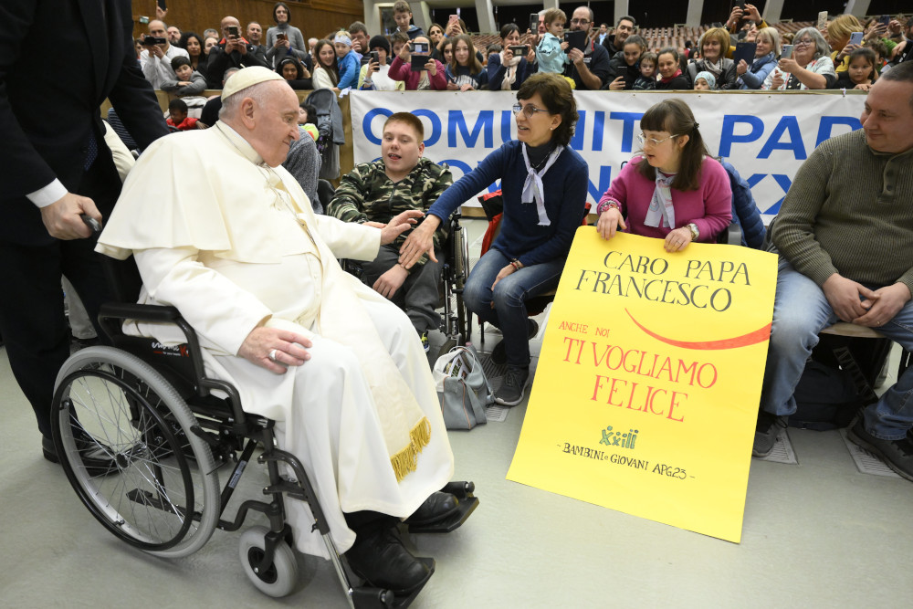 Pope Francis, sitting in a wheelchair, rolls by seated guests in front of a crowd