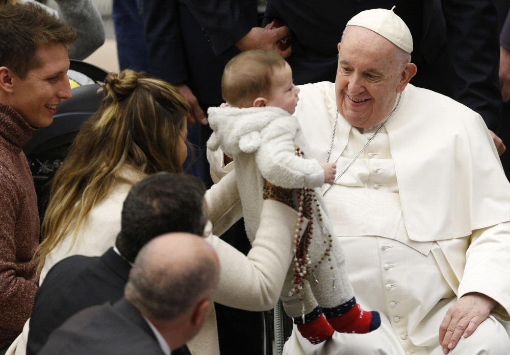 Pope Francis smiles at a baby who is held up towards him by a woman who also clutches many rosaries to the baby's side