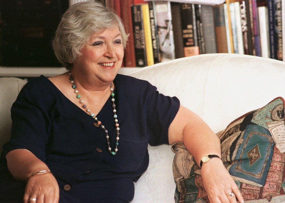 An older white woman wearing a blue dress and a beaded necklace sits on a white couch in front of a bookshelf