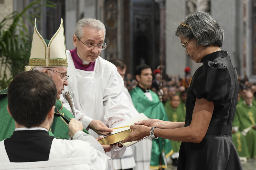 Pope Francis, who is sitting, hands a Bible to a standing woman with short gray hair and a black dress