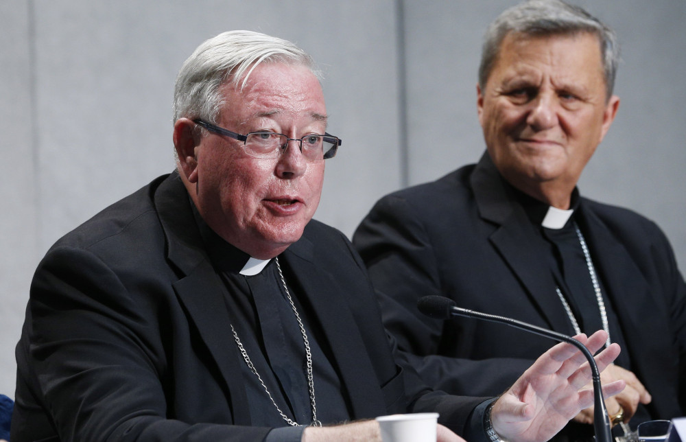 An older white man in a clergy collar and black suit and pectoral cross speaks into a microphone and is sitting next to another similarly dressed older white man